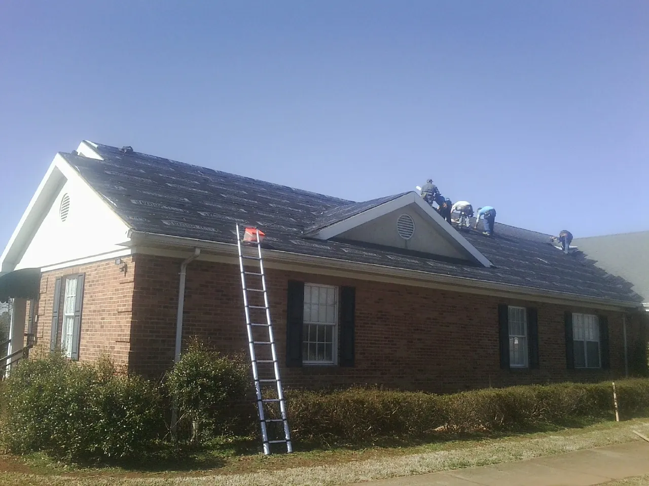 Crew tearing shingles off a brick ranch roof with a ladder set — Metal Roof Repair work by our crew in Moore Haven